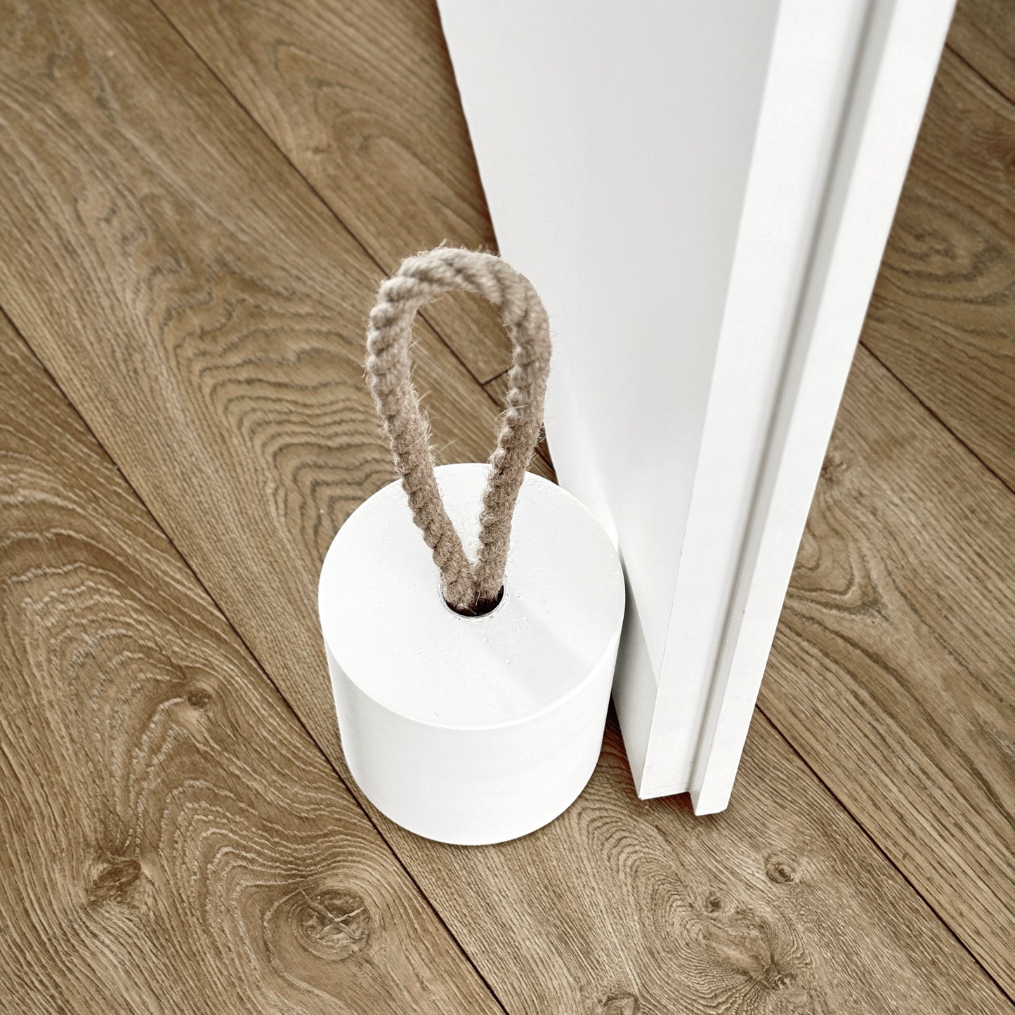 Top-down close-up of an extra-large white concrete door stopper with a cylindrical shape and natural jute handle, positioned against an open interior door in a neutral modern interior. Designed to keep the door open.