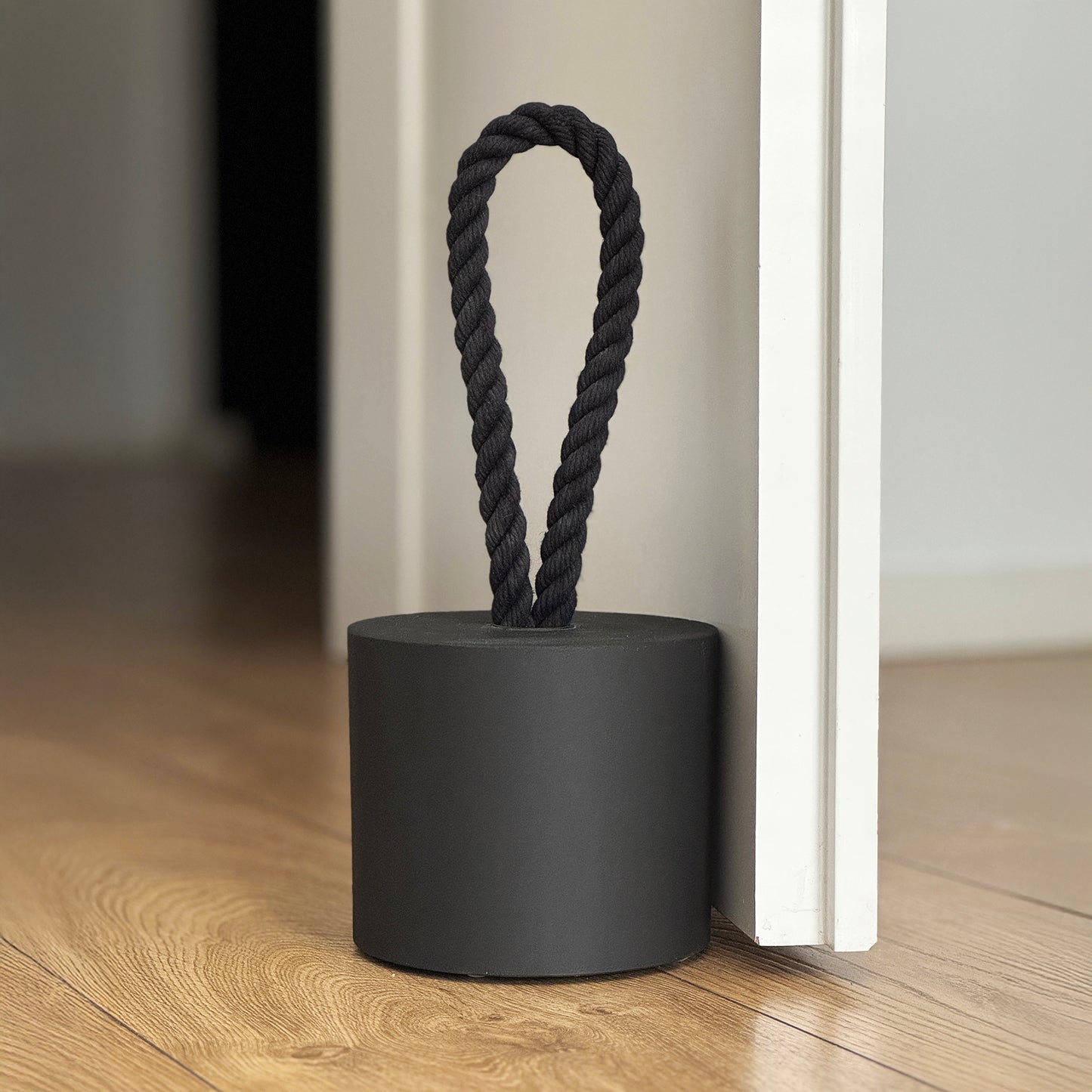 Close-up of an extra-large black concrete door stopper in a cylindrical shape with a black cotton handle, positioned against a door in a neutral modern interior. A sleek, minimalist design, perfect for contemporary door stops in modern spaces.