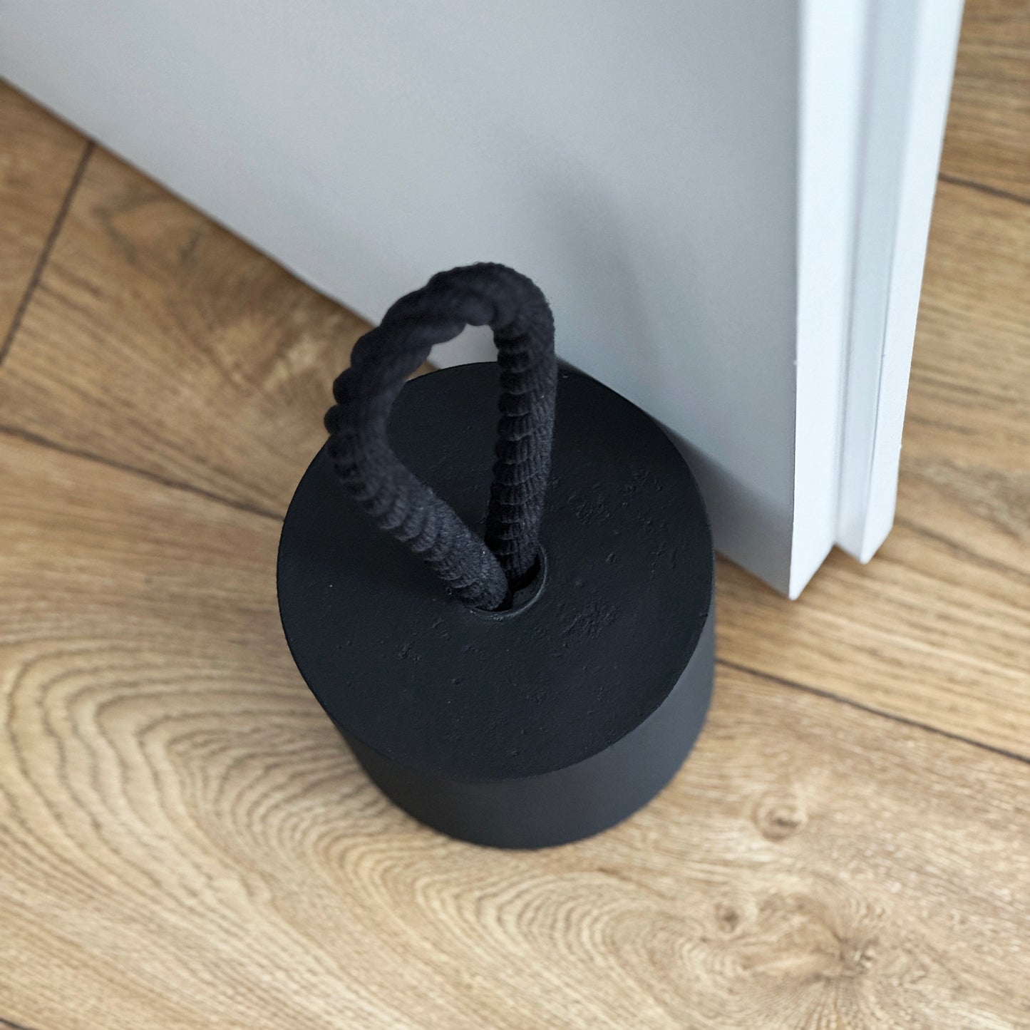 Top-down close-up of an extra-large black concrete door stopper with a cylindrical shape and black cotton handle, positioned against an open interior door in a neutral modern interior. Designed to keep the door open.