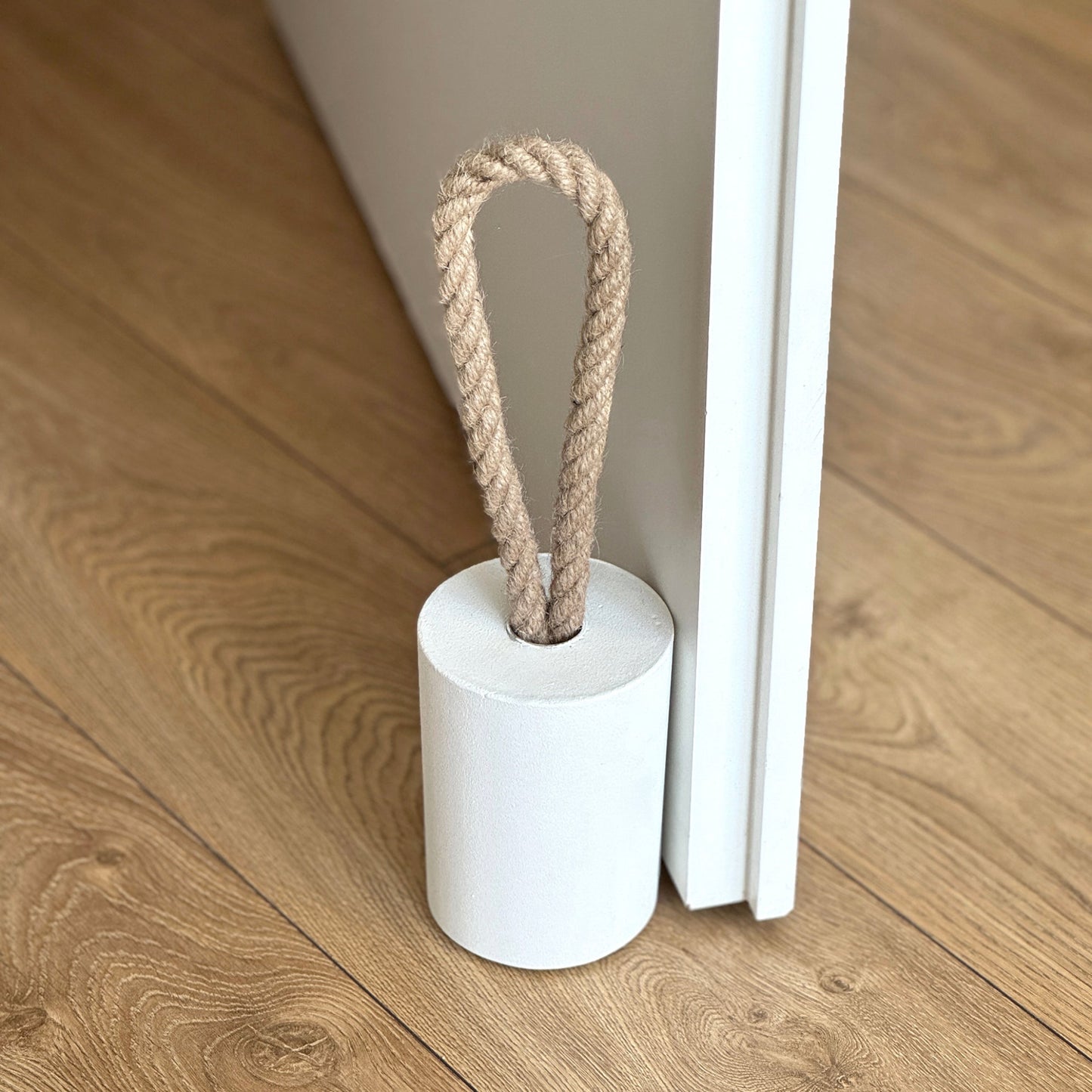 Top side view of a concrete door stop with a natural jute handle in white concrete, placed next to an open white door in a modern interior. The focus is on the door stop and the lower part of the door, with the background softly blurred to highlight the product.