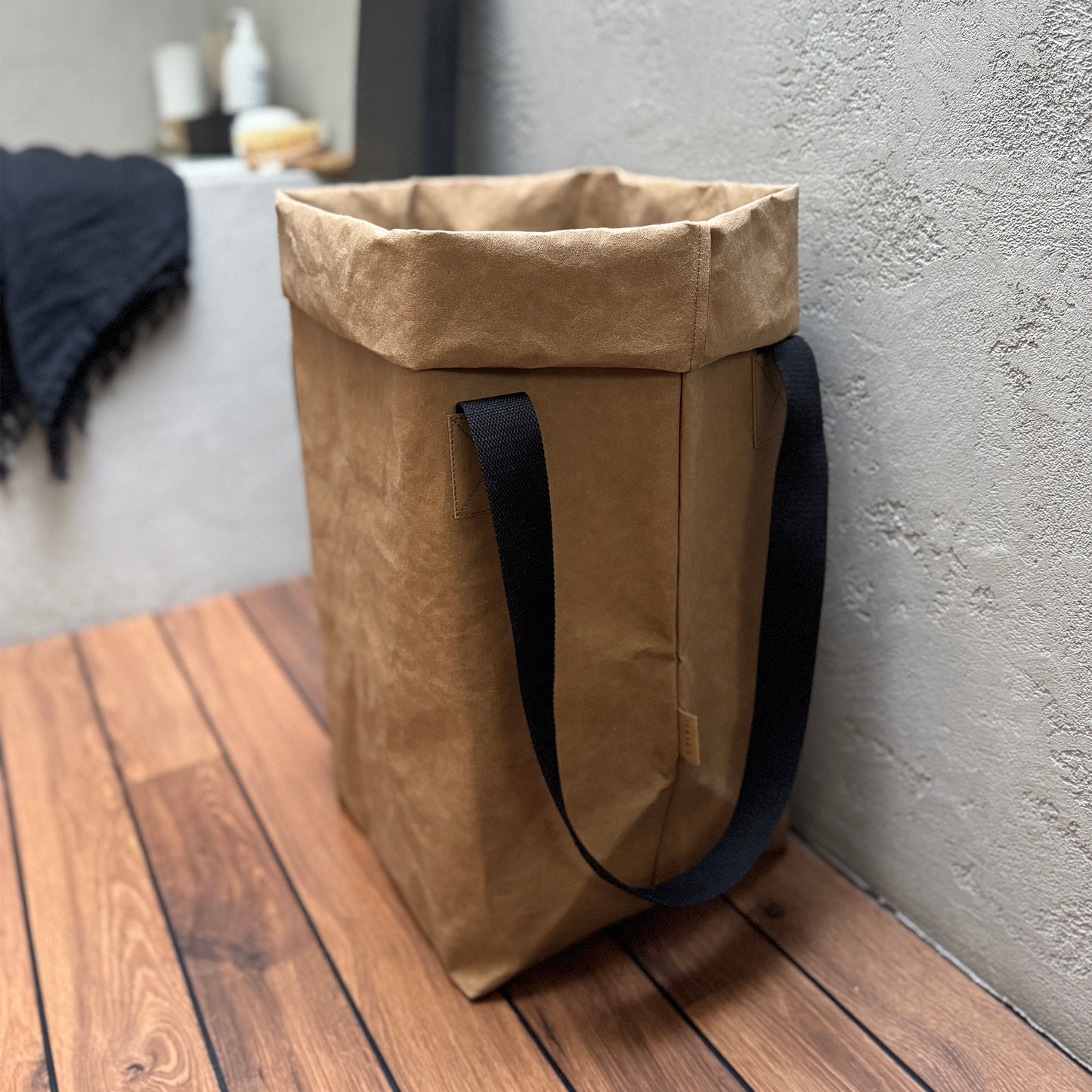 A brown washable paper laundry bag with black cotton handles on a wooden surface with a textured wall in the background.