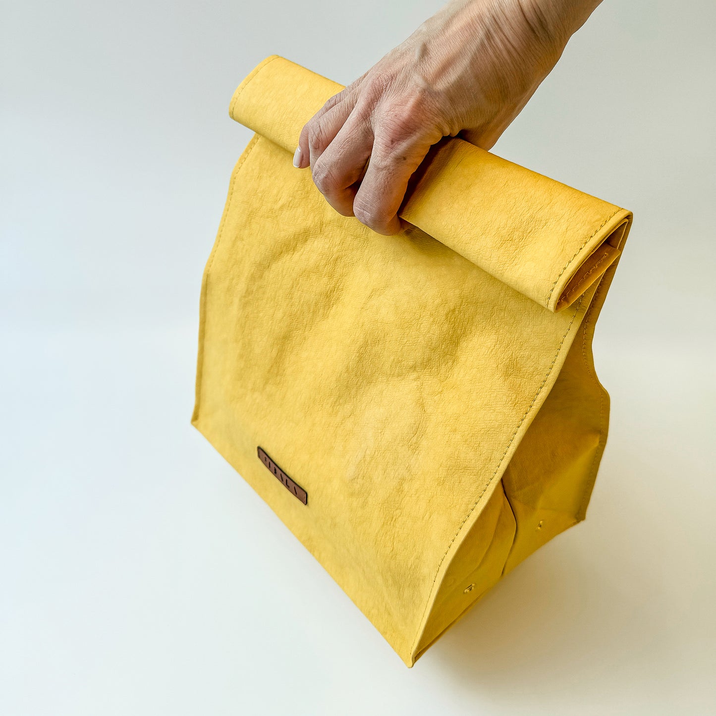 Hand holding a yellow washable paper lunch bag by its folded top, demonstrating lightweight and easy-carry design on white background.