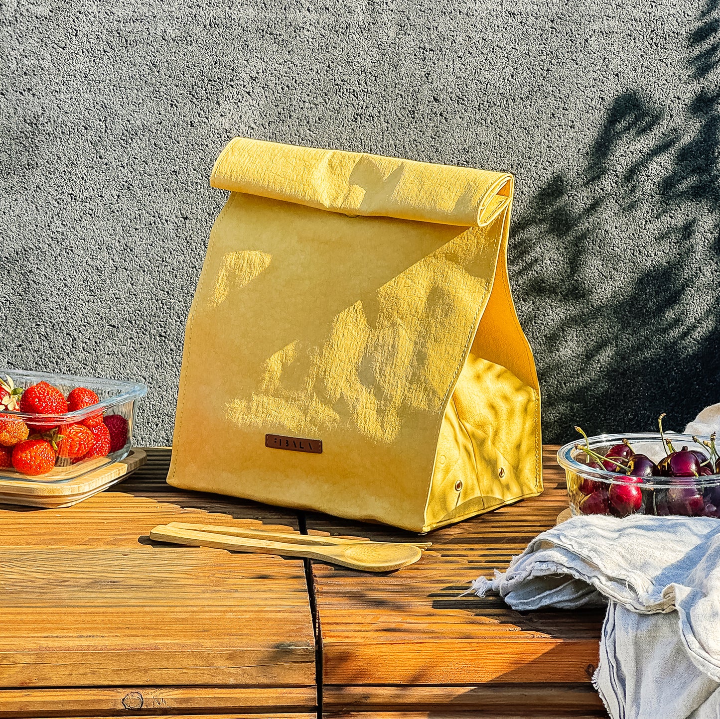 Yellow washable paper lunch bag with snap closure, minimalist eco lunch tote with fresh fruit containers on bench against concrete wall