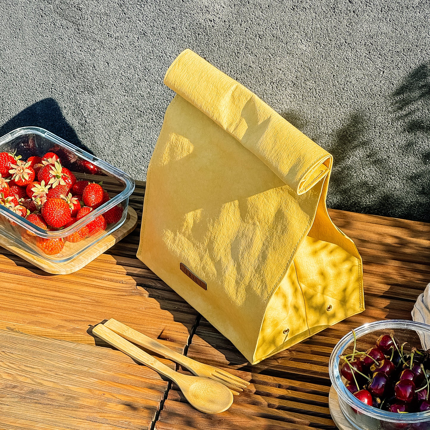 Top-side view of yellow washable paper lunch bag with folded top and metal snap closure, styled with fruit containers on wooden bench and concrete wall backdrop