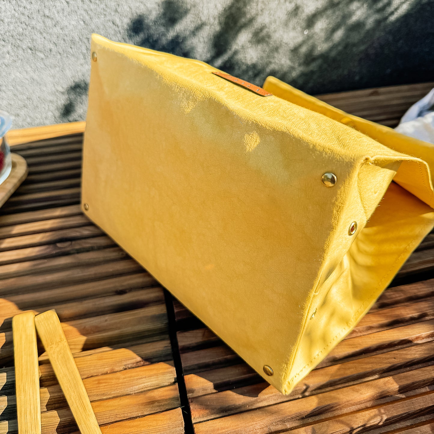 Close-up bottom view of yellow washable paper lunch bag with snap closure, showing metal feet detail, minimalist eco lunch tote on wooden bench with concrete wall