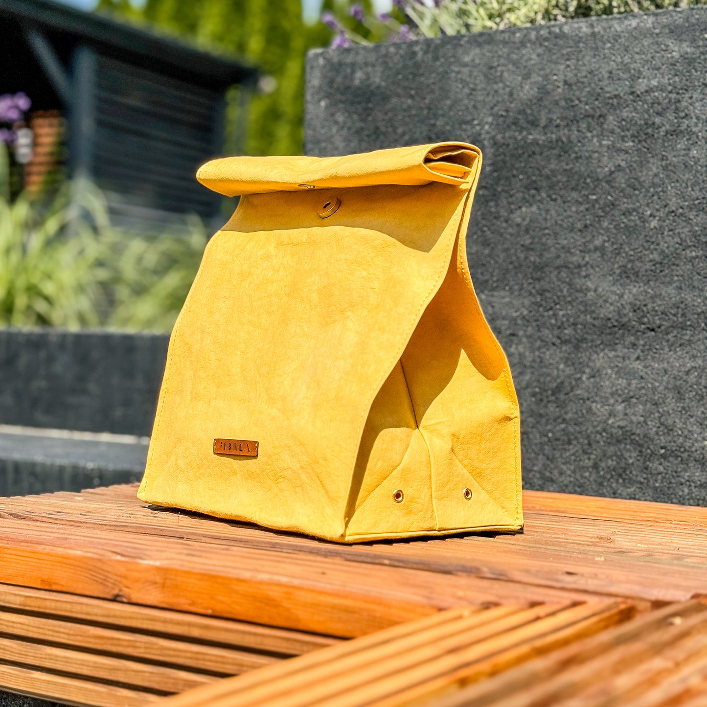 Side view of yellow washable paper lunch bag with folded top and snap closure, standing on wooden bench with concrete planters and garden background.