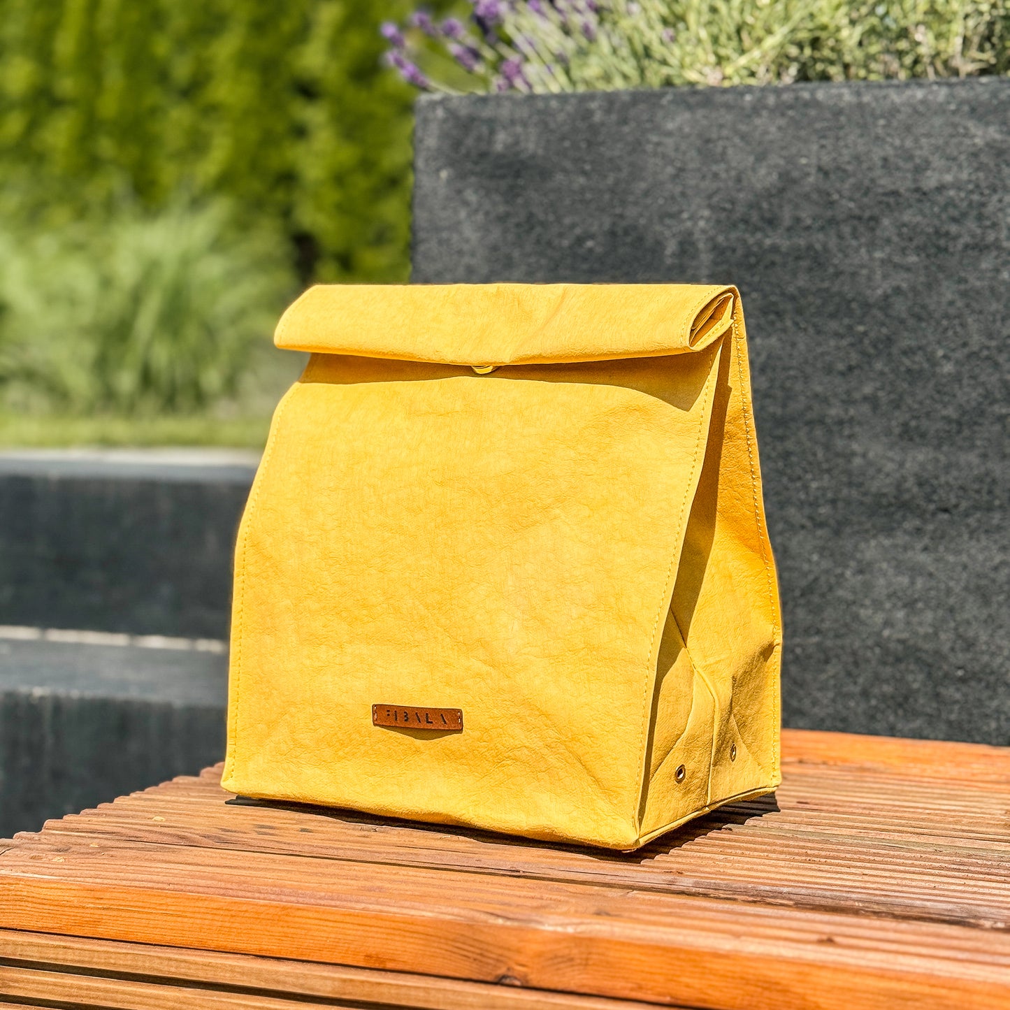 Front view of yellow washable paper lunch bag with folded top and snap closure, standing on wooden bench with concrete planters and garden background.