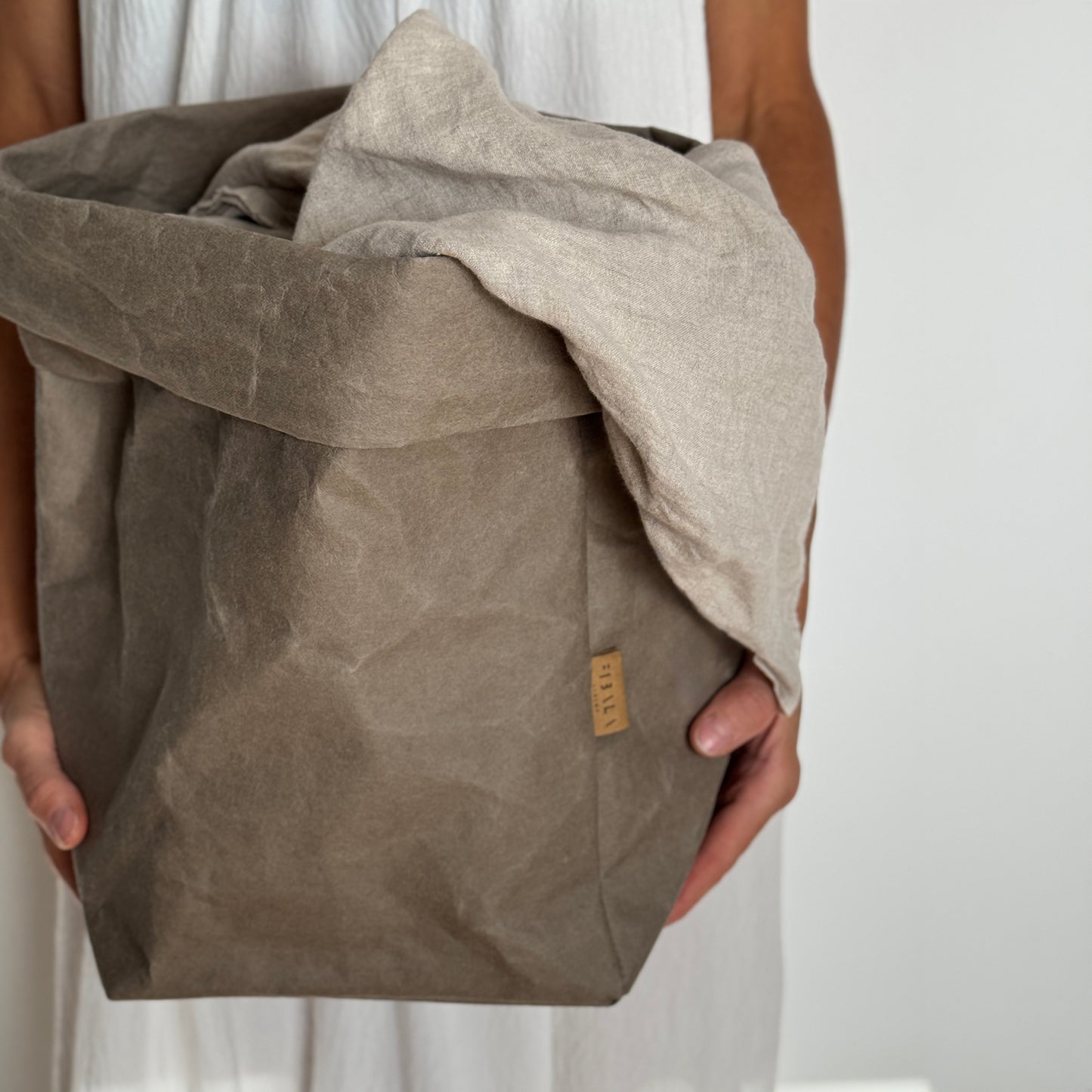 Individual grasping an oversized chino brown washable paper bag, draped with a beige linen cloth, in front of a white backdrop | Sustainable paper bag