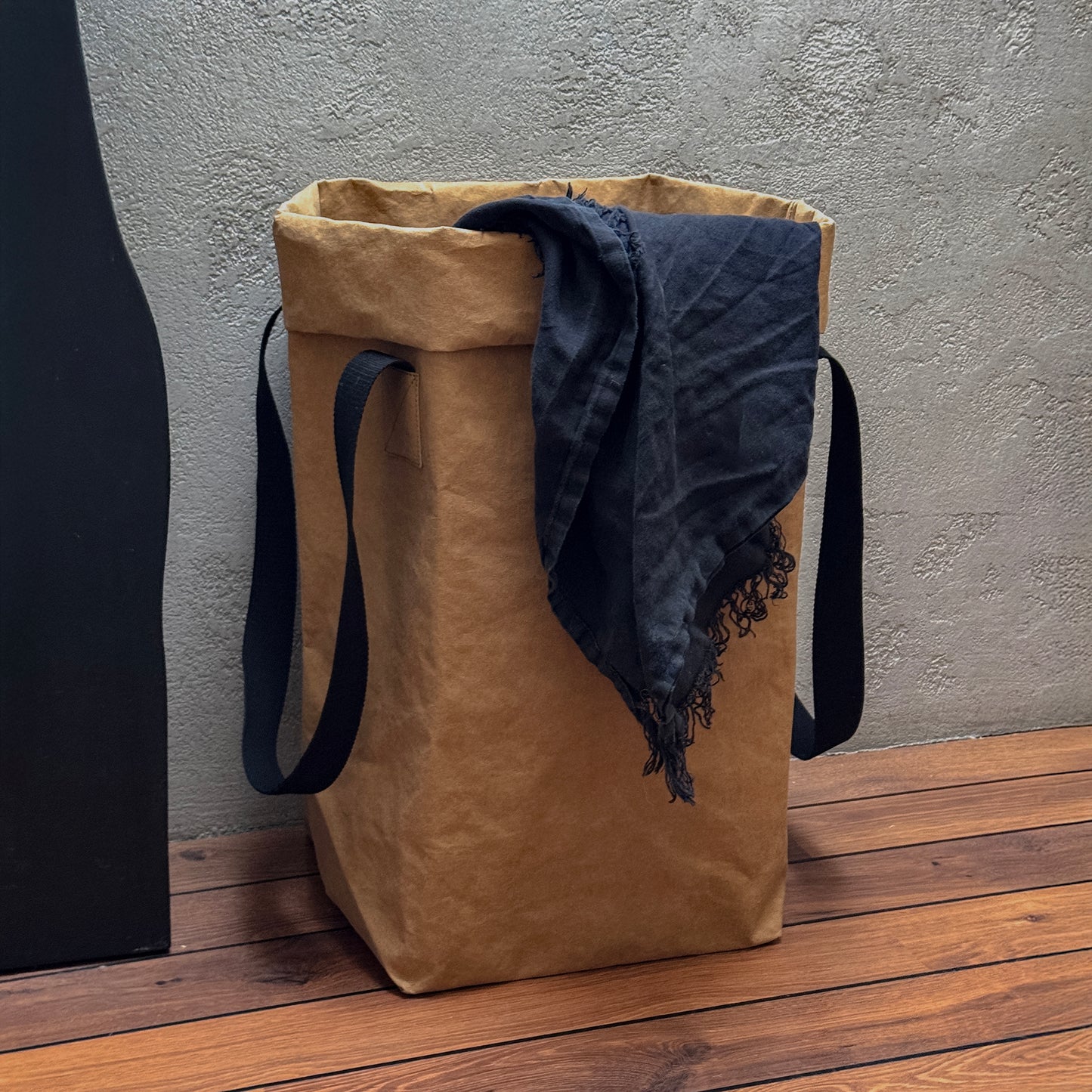A brown washable paper laundry bag with black cotton handles on a wooden surface with a textured wall behind.