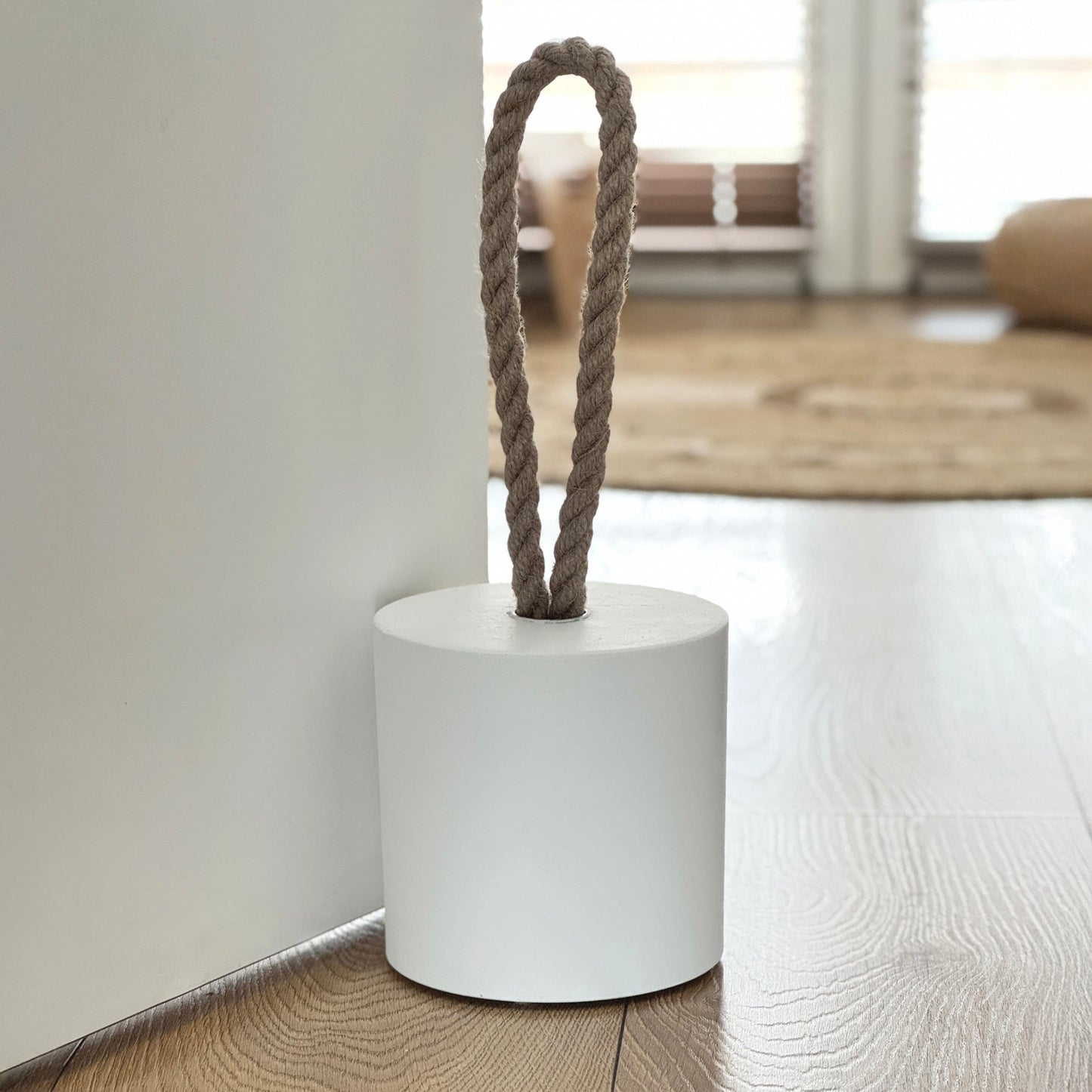 Front view of an extra-large white concrete door stopper with a cylindrical shape and natural jute handle placed against a door in a neutral modern interior. Sleek and minimalist, ideal for contemporary spaces and suitable for outdoor use.