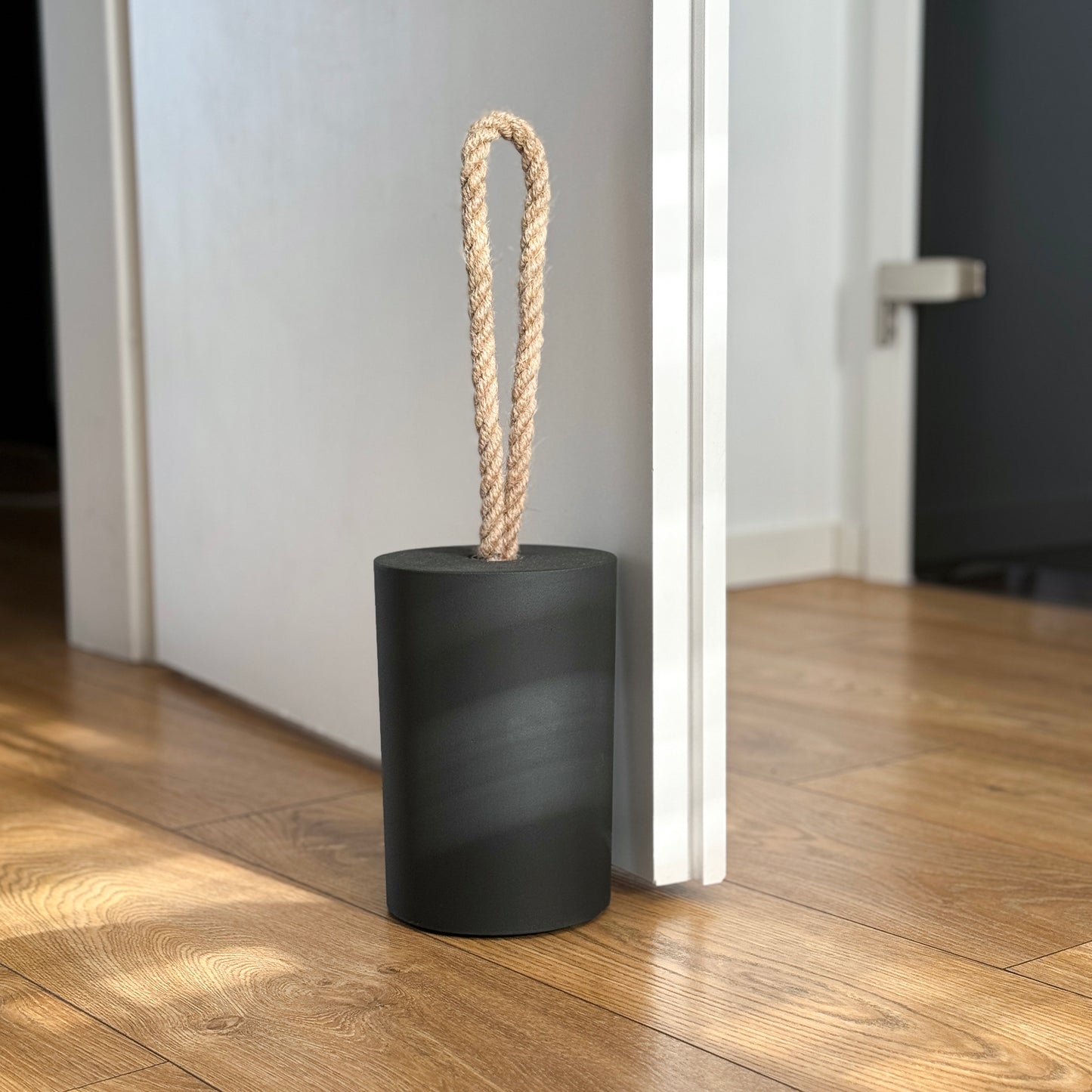 Extra large black concrete door stop with a loop-style natural jute rope handle, positioned beside an open door in a modern interior. The close-up highlights the textured concrete surface and twisted rope detail, designed for easy lifting. A bold, functional accent for modern, minimalist spaces and stylish interior.