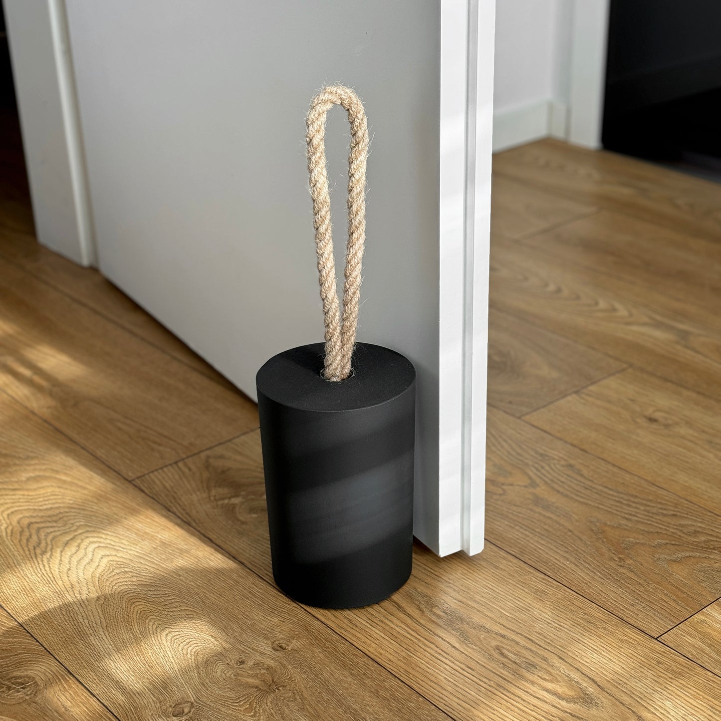 Extra large black concrete door stop with a loop-style natural jute rope handle, positioned beside an open door in a modern interior. The top-side highlights the textured concrete surface and twisted rope detail, designed for easy lifting. A bold, functional accent for modern, minimalist spaces and stylish interior.