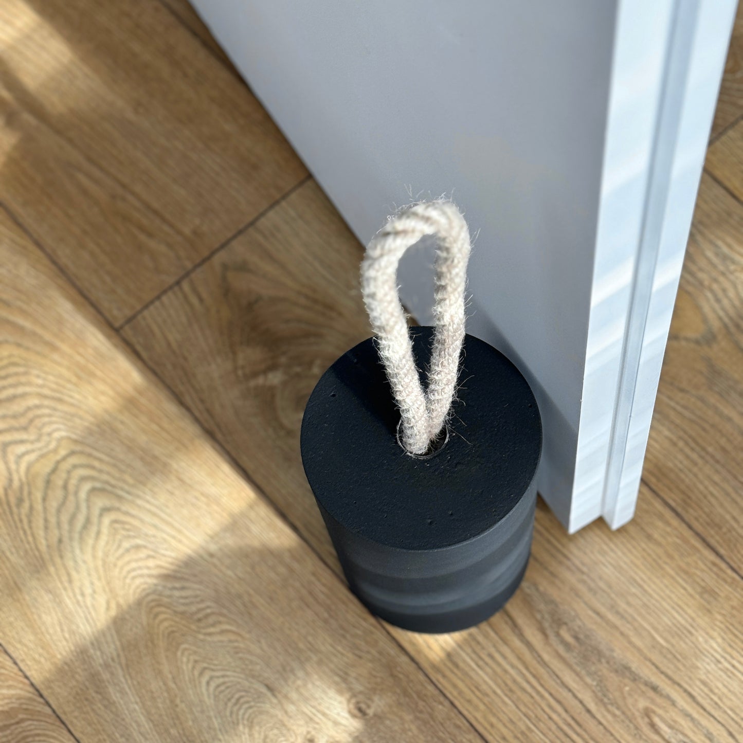 Top-down close-up of an extra large black concrete door stopper with a cylindrical shape and natural jute handle, positioned against an open interior door in a neutral modern interior. Designed to keep the door open.