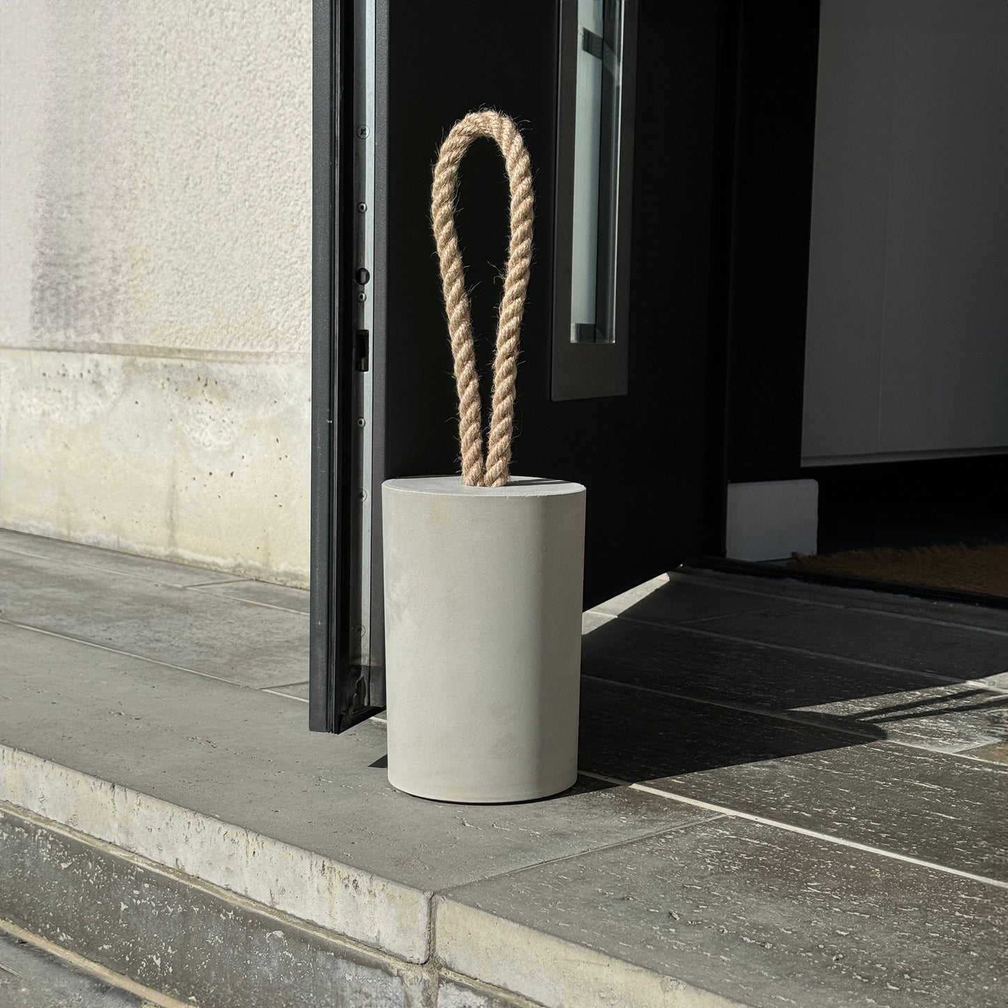 Extra large grey concrete door stop with a loop-style natural jute rope handle, positioned beside an open exterior door. The close-up highlights the textured concrete surface and twisted rope detail, designed for easy lifting. A bold, functional accent for modern, minimalist spaces and stylish exterior.