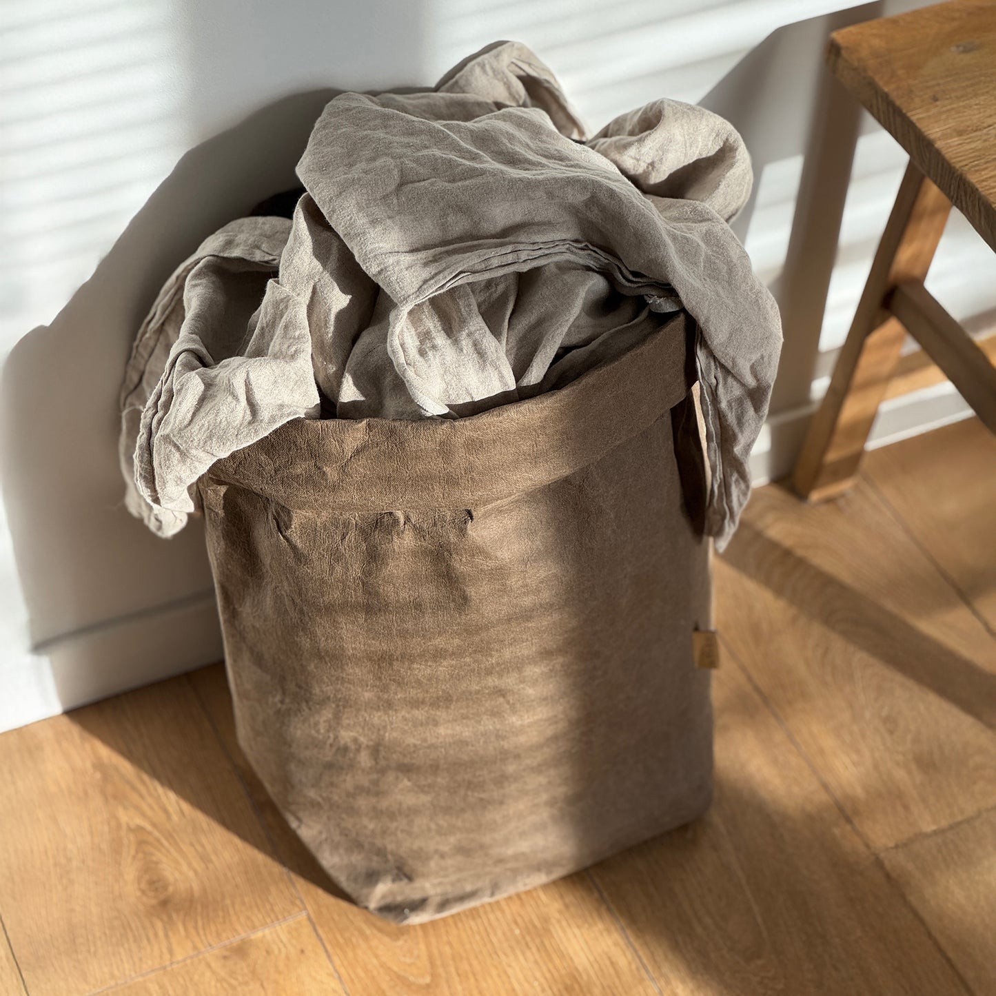 Top view of XXL cocoa brown washable paper bag filled with linen throw, on wooden floor, white wall background | Eco-friendly, versatile storage solution