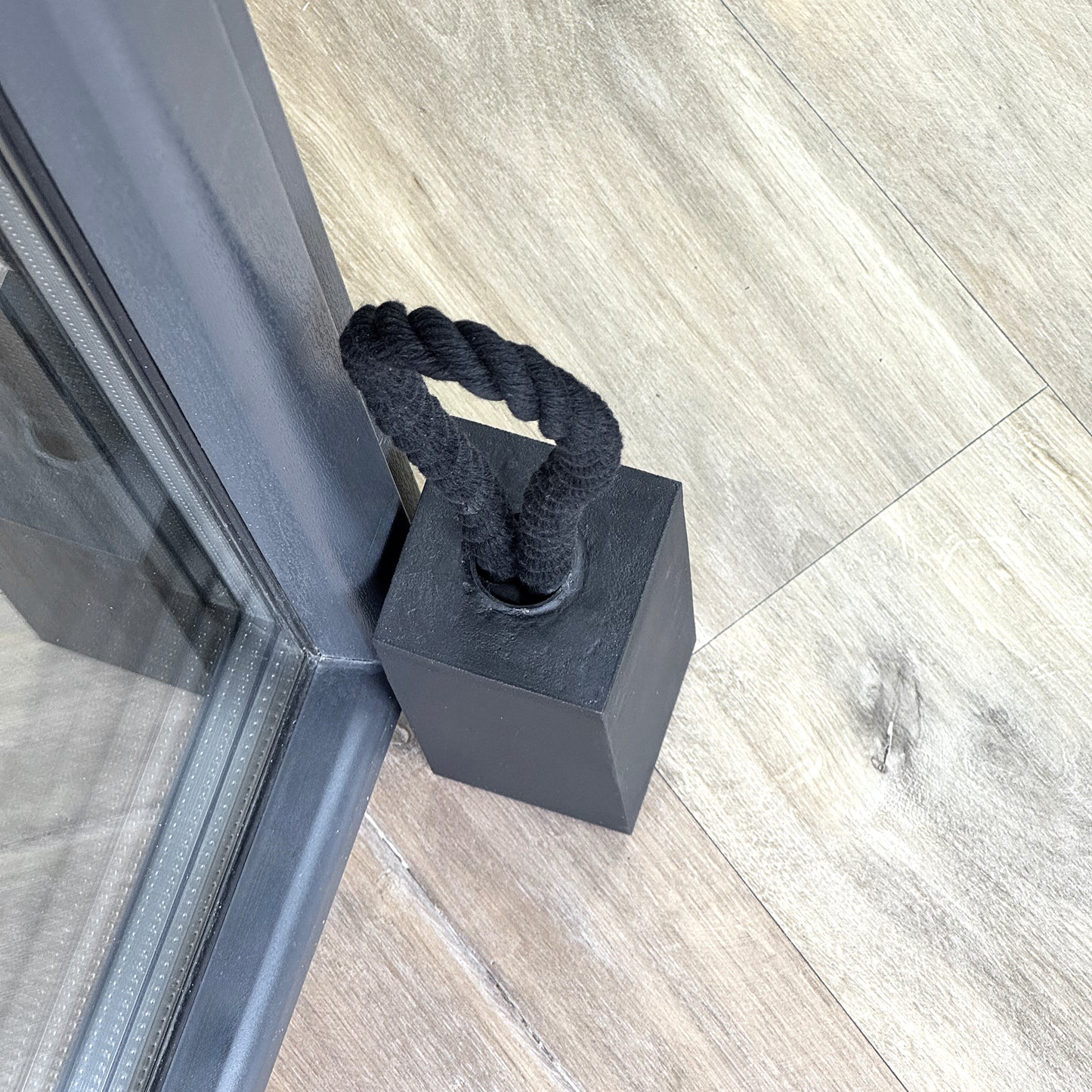 Black concrete door stop in rectangular shape with a black cotton rope handle placed next to a glass door on a wooden floor. Close-up top view.