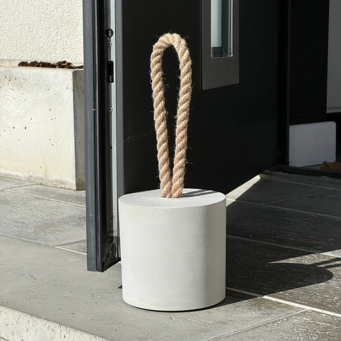 Close-up of an extra-large grey concrete door stopper in a cylindrical shape with a jute handle, holding an exterior door open in a modern outdoor setting. A minimalist, modern door stop designed for outdoor use.