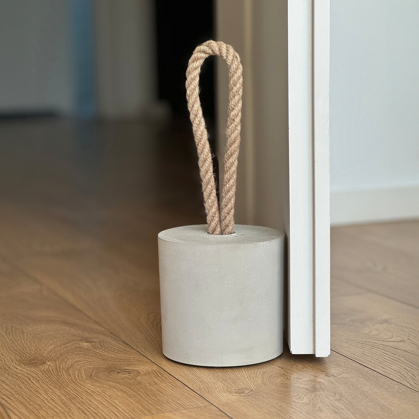 Front view of an extra-large grey concrete door stopper with a cylindrical shape and jute handle, placed against a door in a neutral modern interior. Sleek and minimalist, ideal for contemporary spaces and suitable for outdoor use.