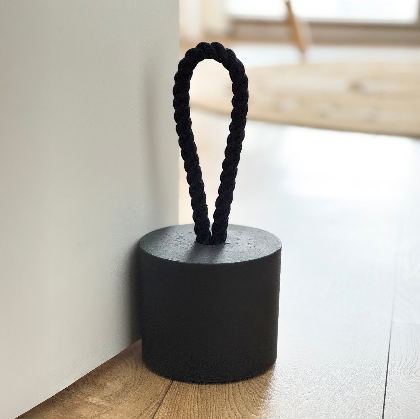 extra-large black concrete door stop in a cylindrical shape with a black cotton handle, positioned against a door in a neutral modern interior.