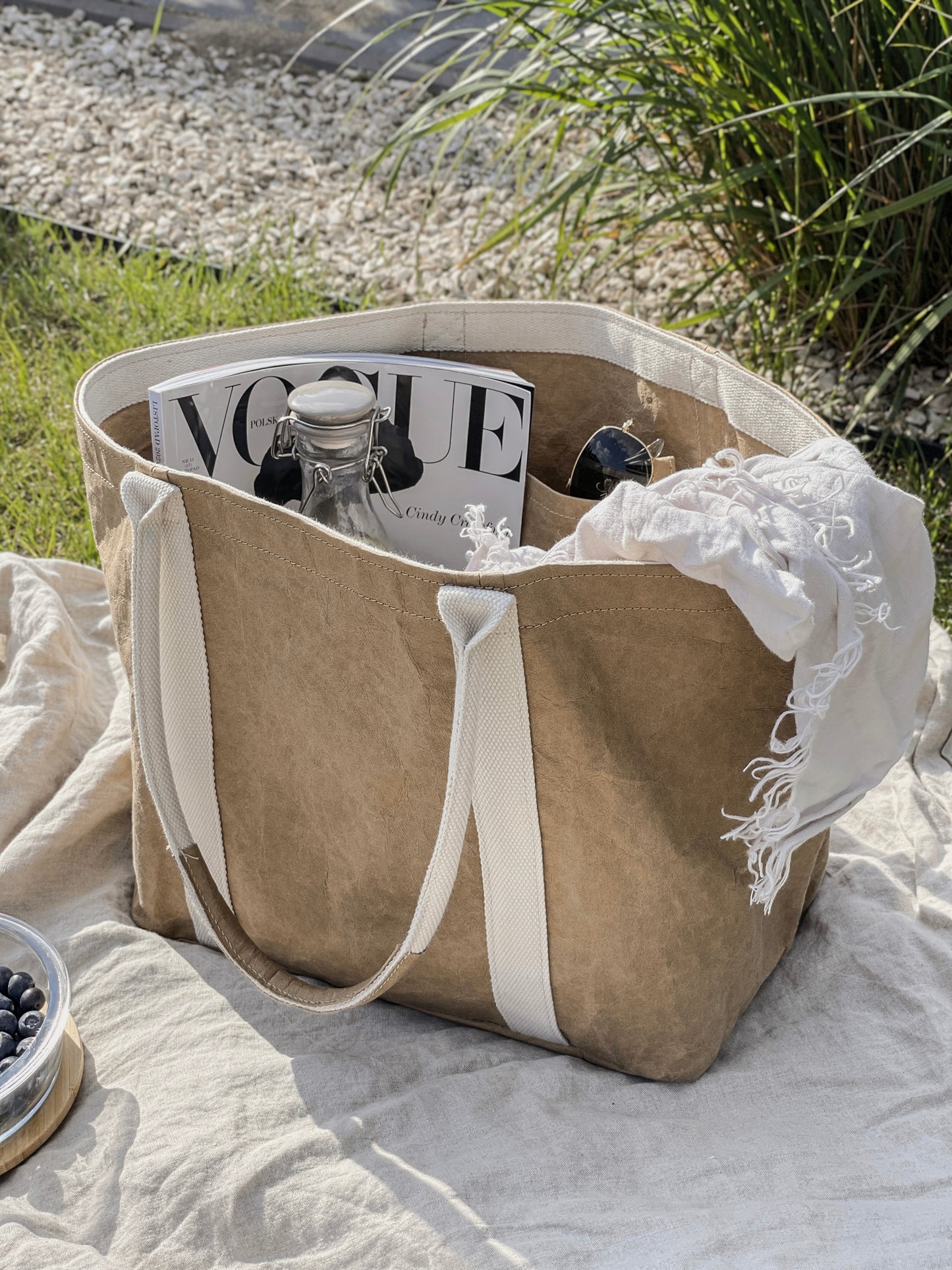 Brown washable paper tote bag at outdoor picnic on linen blanket, filled with Vogue magazine, glass bottle, sunglasses and white linen scarf