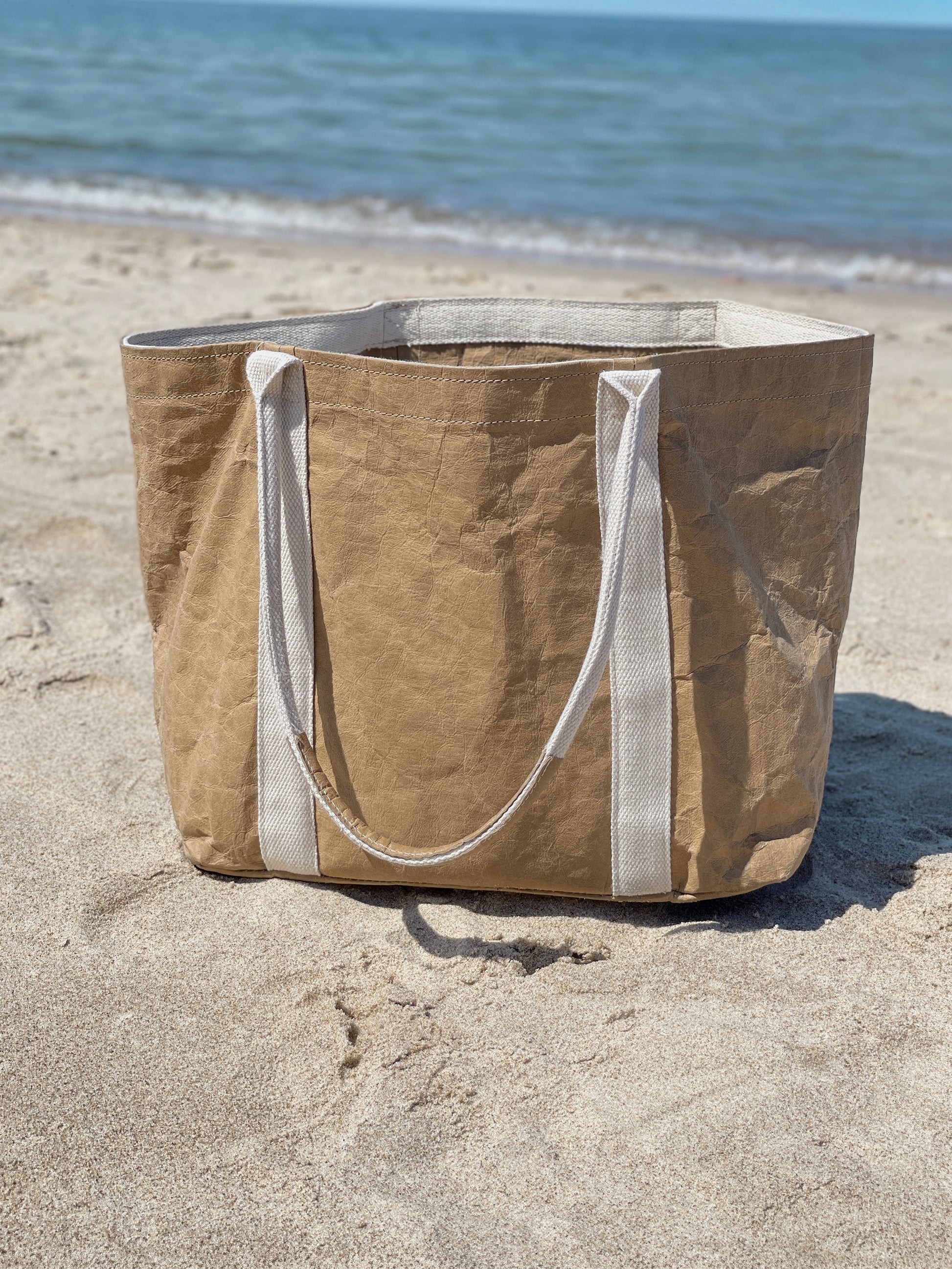 Brown washable paper tote bag standing on sand, structured flat bottom, ocean in background