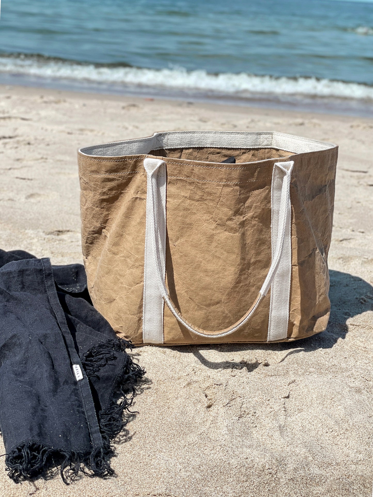 Brown washable paper tote bag on beach sand beside a dark beach towel, sea in background