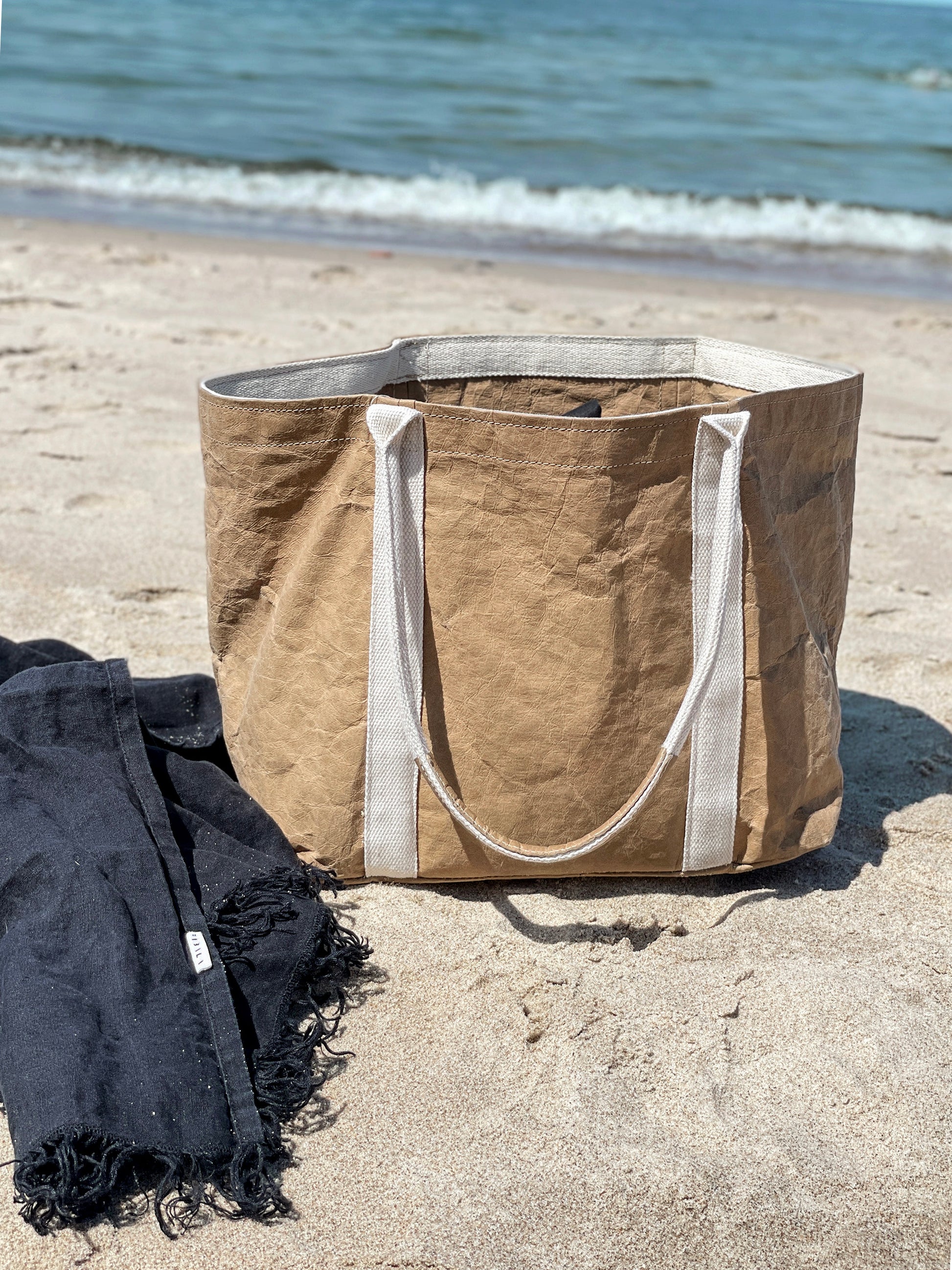 Brown washable paper tote bag on beach sand beside a dark beach towel, sea in background