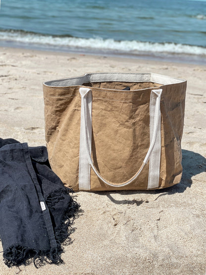Brown washable paper tote bag on beach sand beside a dark beach towel, sea in background