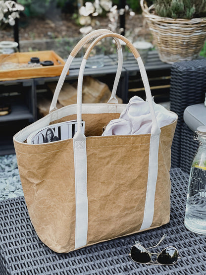 Brown washable paper tote bag on outdoor table, Vogue magazine inside, sunglasses and glass bottle beside it, garden setting