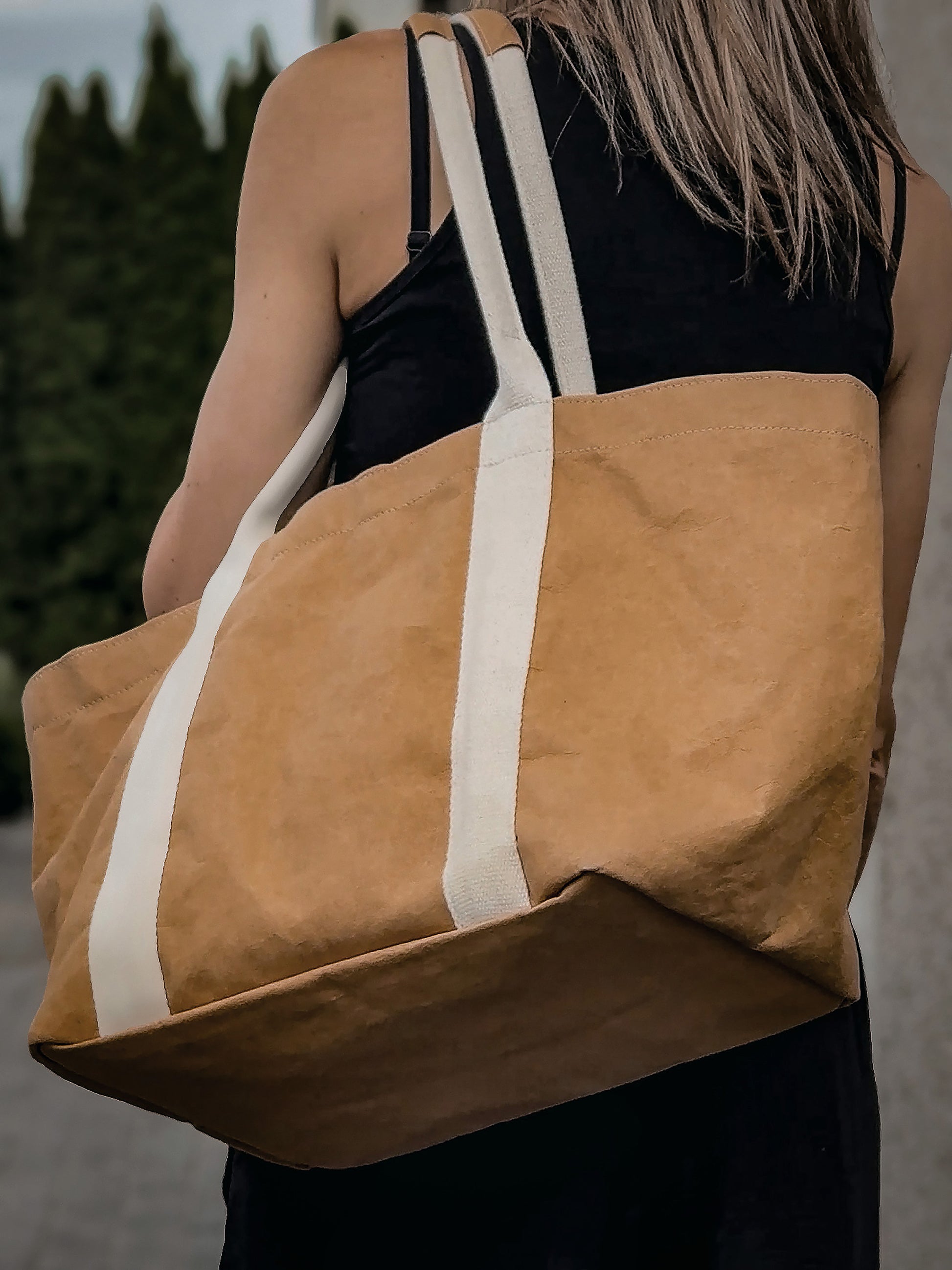 Brown washable paper tote bag worn on shoulder from behind, close-up, black top, outdoor setting
