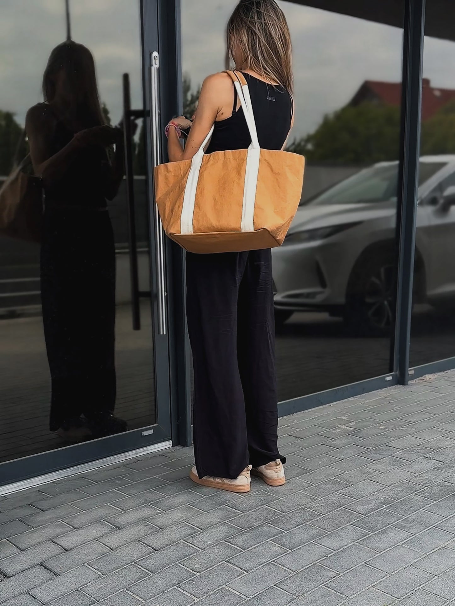 Woman from behind carrying brown washable paper tote on shoulder, black outfit, standing by glass entrance, urban street reflection
