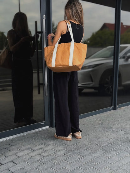 Woman from behind carrying brown washable paper tote on shoulder, black outfit, standing by glass entrance, urban street reflection