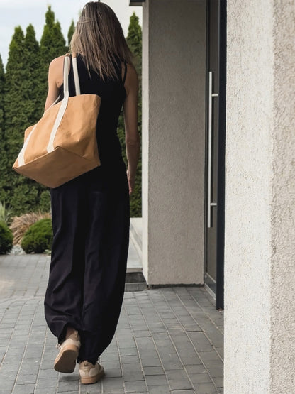 Woman walking away on paved path, black wide-leg jumpsuit, brown washable paper tote on shoulder, garden and building background