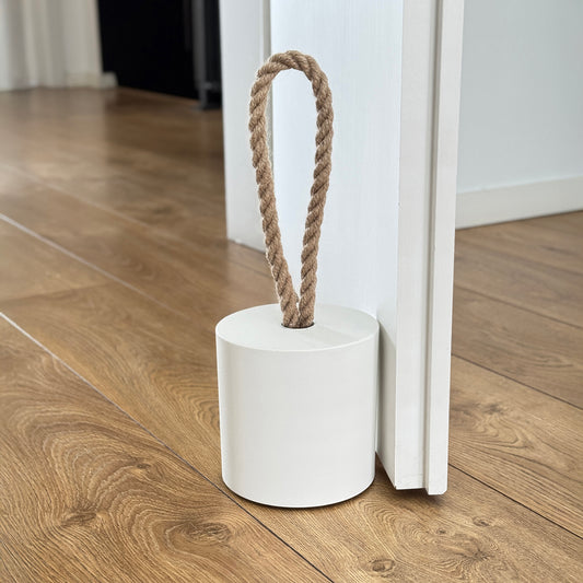 Front view of an extra-large white concrete door stopper with a cylindrical shape and jute handle, placed against a door in a neutral modern interior. Sleek and minimalist, ideal for contemporary spaces and suitable for outdoor use.