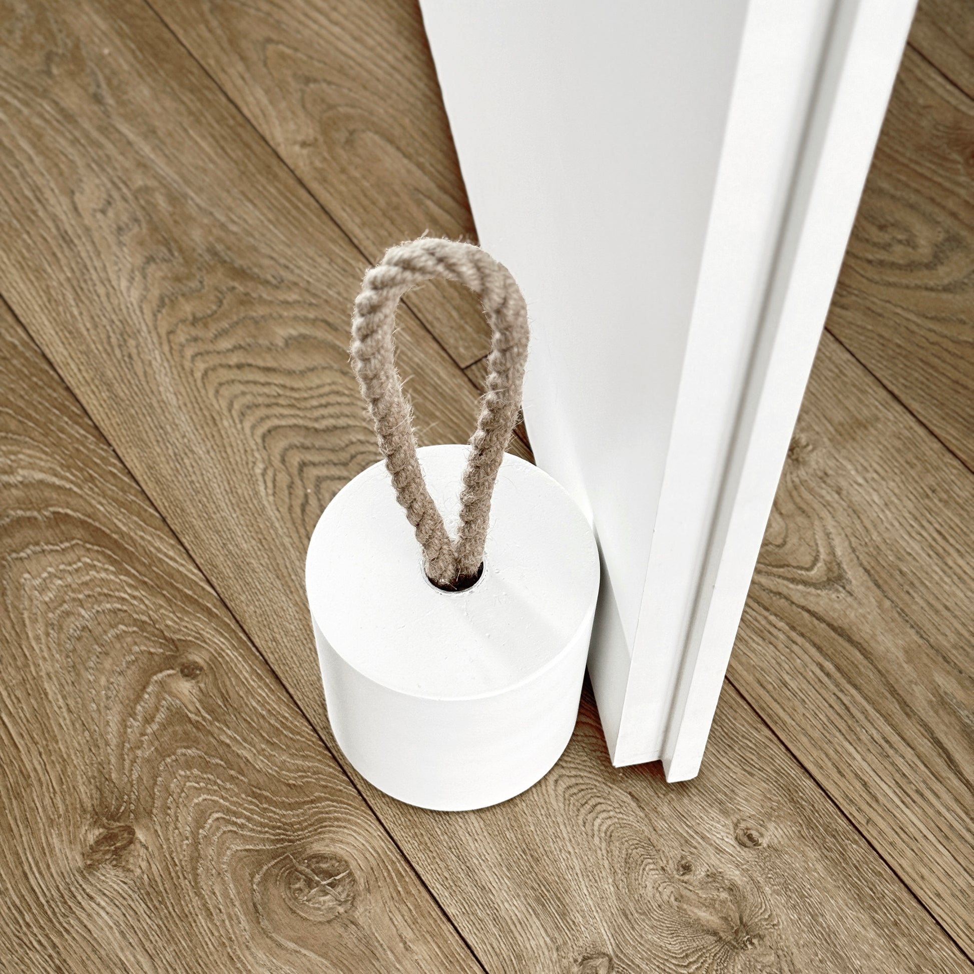 Top-down close-up of an extra-large white concrete door stopper with a cylindrical shape and natural jute handle, positioned against an open interior door in a neutral modern interior. Designed to keep the door open.