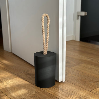 Side view of a concrete door stop with a natural jute handle in black, placed next to an open white door in a modern interior. The focus is on the door stop and the lower part of the door, with the background softly blurred to highlight the product.