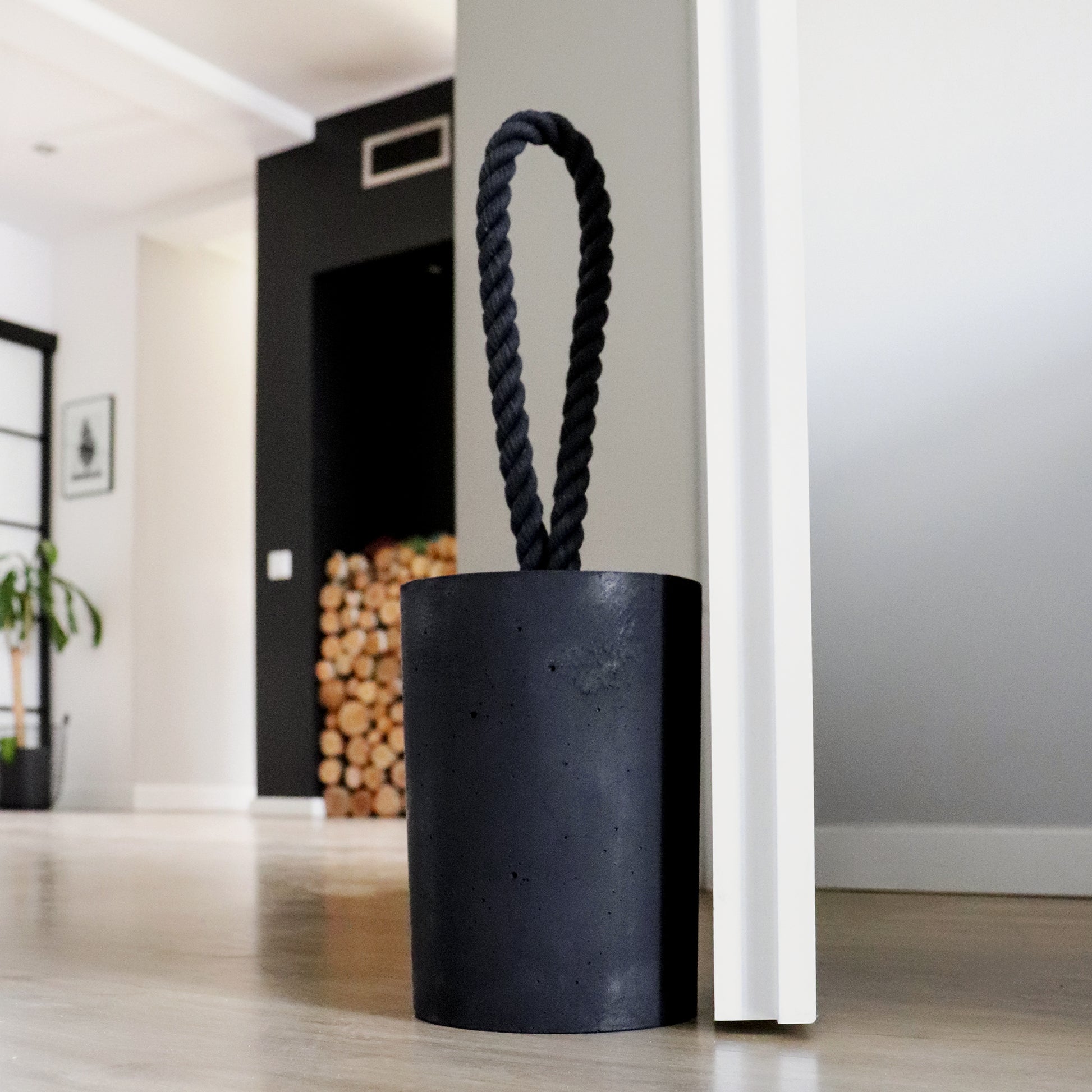 Extra large black concrete door stop with a loop-style black cotton rope handle, positioned beside an open door in a modern interior. The close-up highlights the textured concrete surface and twisted rope detail, designed for easy lifting. A bold, functional accent for modern, minimalist spaces and stylish interiors or exteriors.