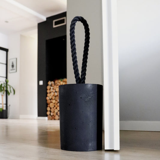 Extra large black concrete door stop with a loop-style black cotton rope handle, positioned beside an open door in a modern interior. The close-up highlights the textured concrete surface and twisted rope detail, designed for easy lifting. A bold, functional accent for modern, minimalist spaces and stylish interiors or exteriors.