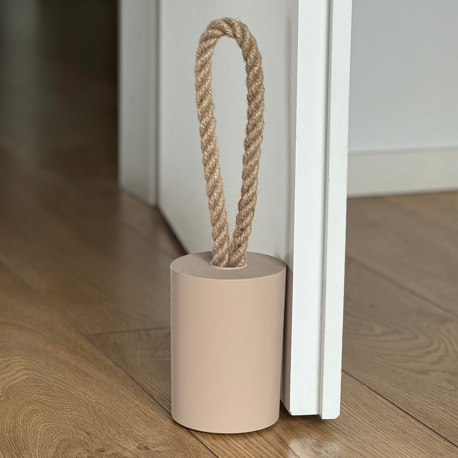 Top side view of a concrete door stop with a natural jute handle in earthy Classic Beige, placed next to an open white door in a modern interior. The focus is on the door stop and the lower part of the door, with the background softly blurred to highlight the product.