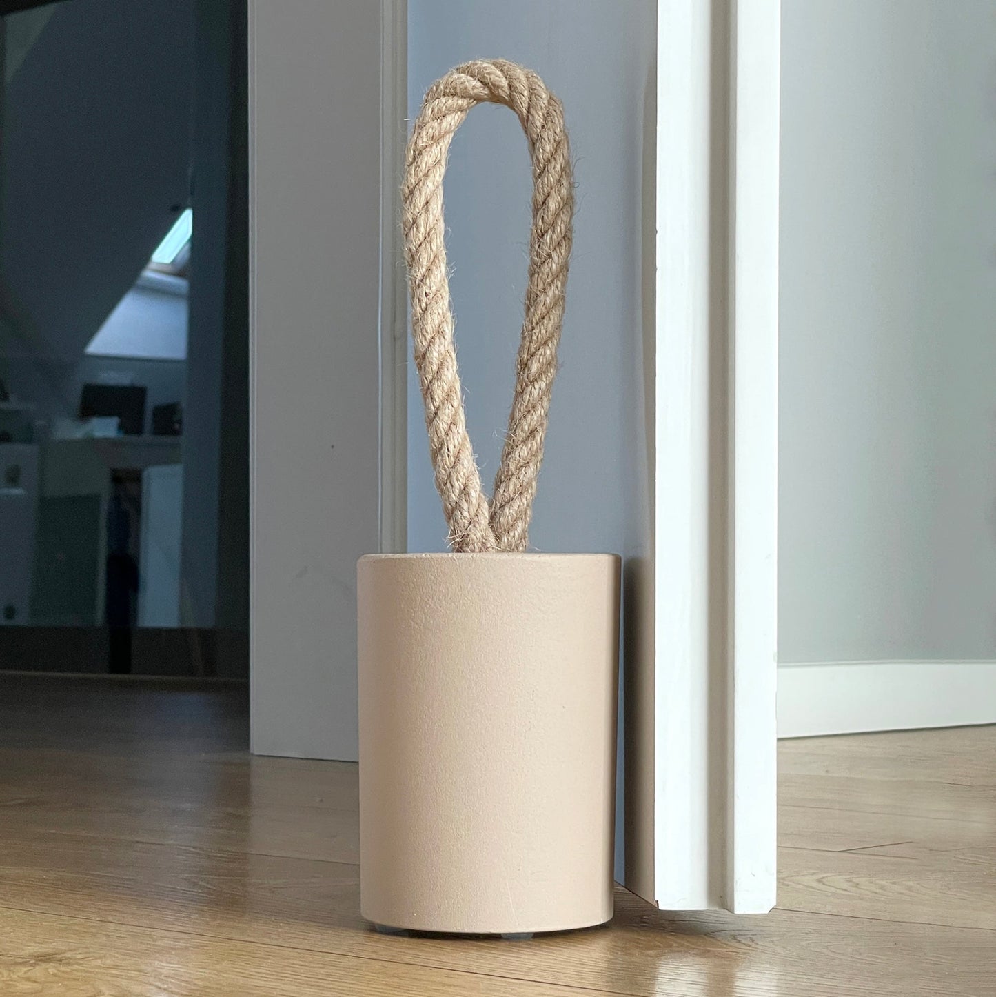 Front view of a concrete door stop with a natural jute handle in earthy Classic Beige, placed next to an open white door in a modern interior. The focus is on the door stop and the lower part of the door, with the background softly blurred to highlight the product.