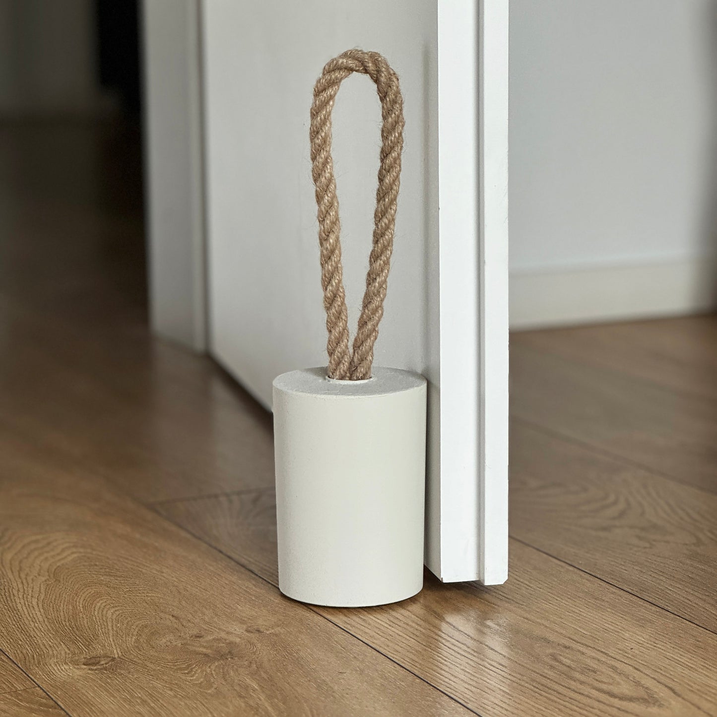 Front view of a concrete door stop with a natural jute handle in earthy Linen White, placed next to an open white door in a modern interior. The focus is on the door stop and the lower part of the door, with the background softly blurred to highlight the product.
