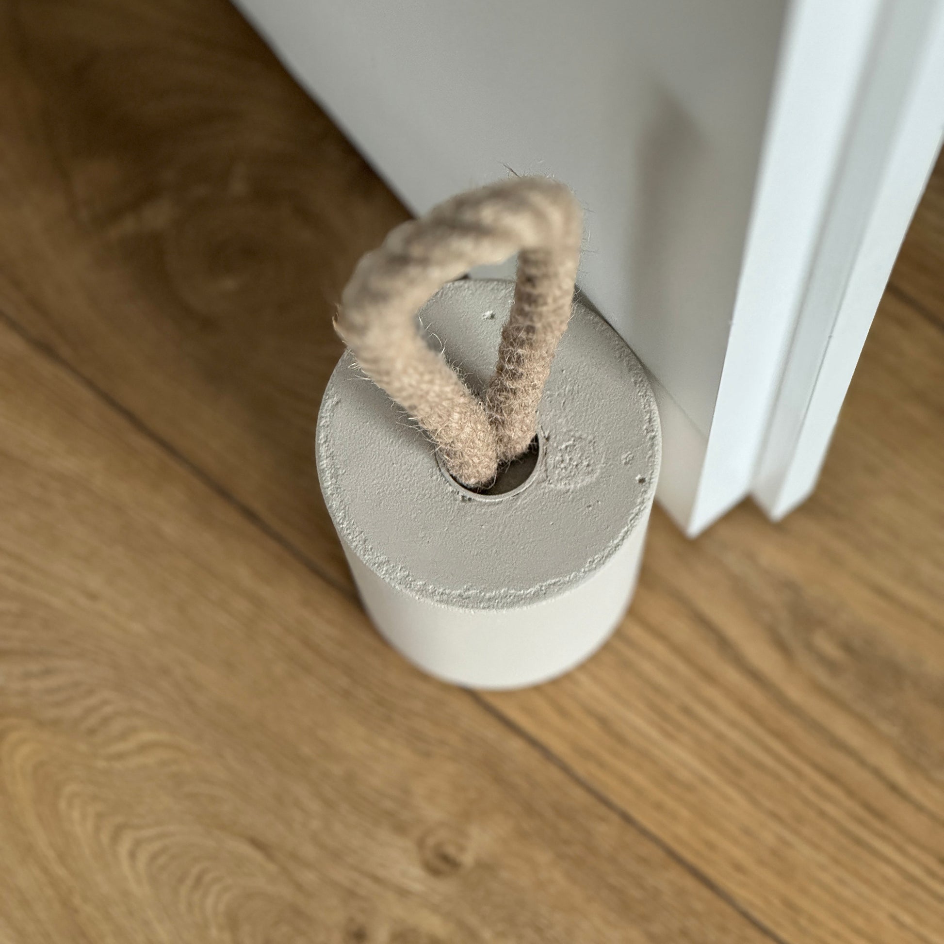 Top-down close-up of a concrete door stop in earthy Pure Beige, featuring a natural jute handle secured with a metal ring. Positioned next to a door, the image highlights the handle detail and showcases a clean, modern aesthetic.