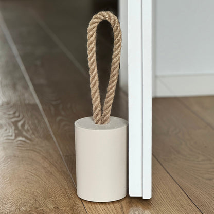 Top side view of a concrete door stop with a natural jute handle in earthy Pure Beige, placed next to an open white door in a modern interior. The focus is on the door stop and the lower part of the door, with the background softly blurred to highlight the product.