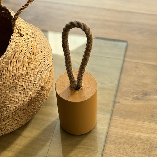 Top view of a concrete door stop with a natural jute handle in earthy Sun Gold Yellow, bathed in warm golden hour light. Positioned on the floor near a handwoven rattan basket, with sunlight streaming from the background to create a cozy, natural ambiance.