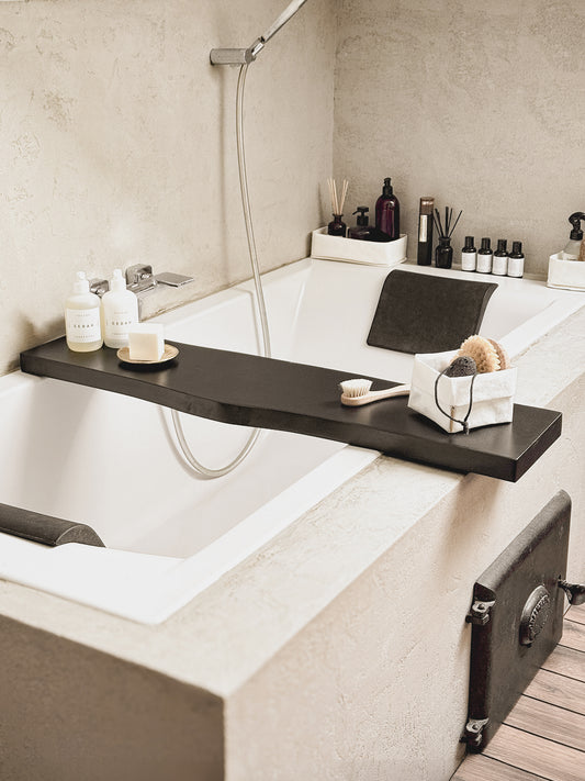 Black live edge bath caddy tray across a built-in tub with skincare, a brush and a small storage bag
