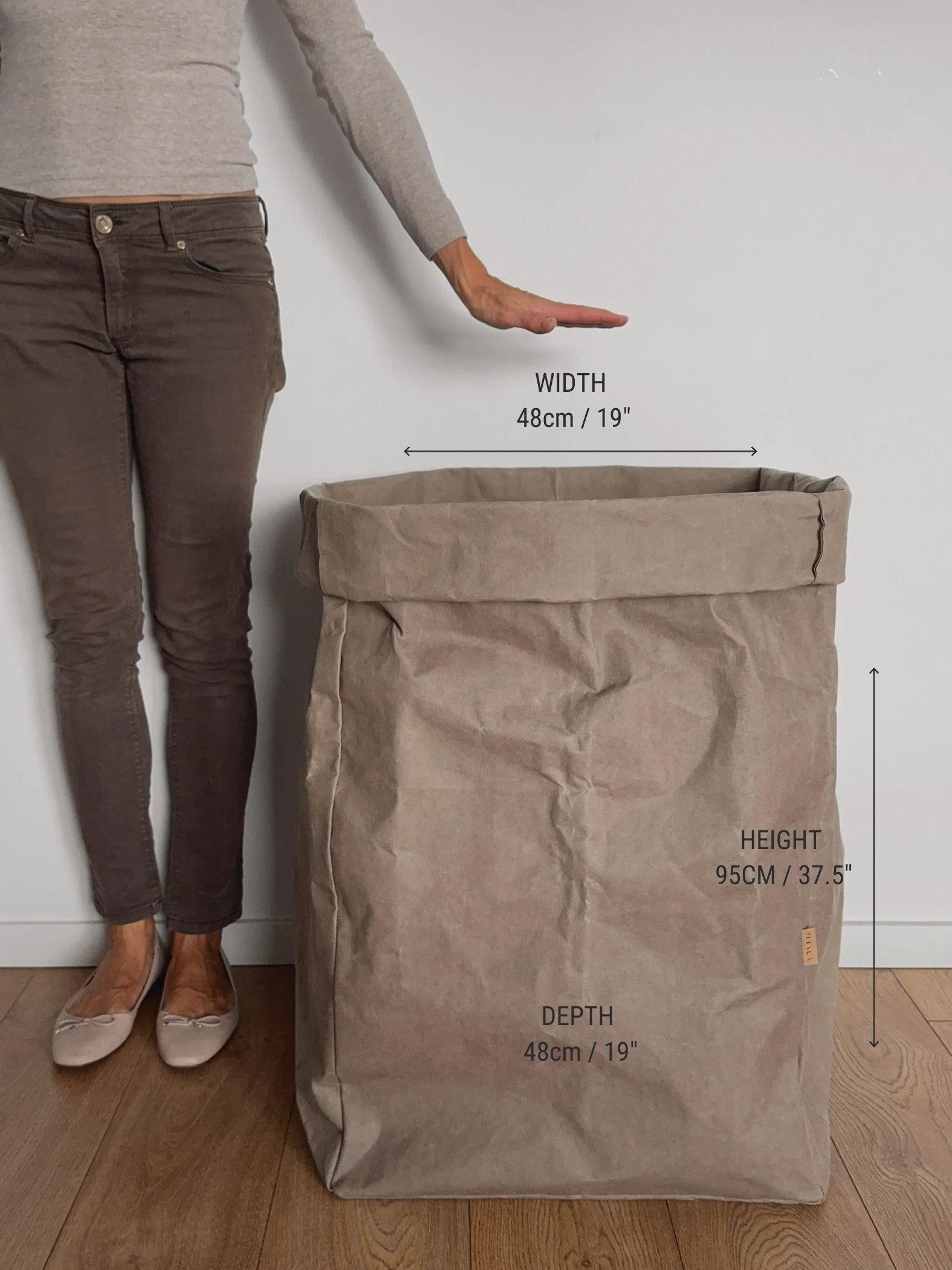 A Giant washable paper storage bag in a natural beige color stands on a wooden floor against a white wall. Showcasing the bag measurements. 