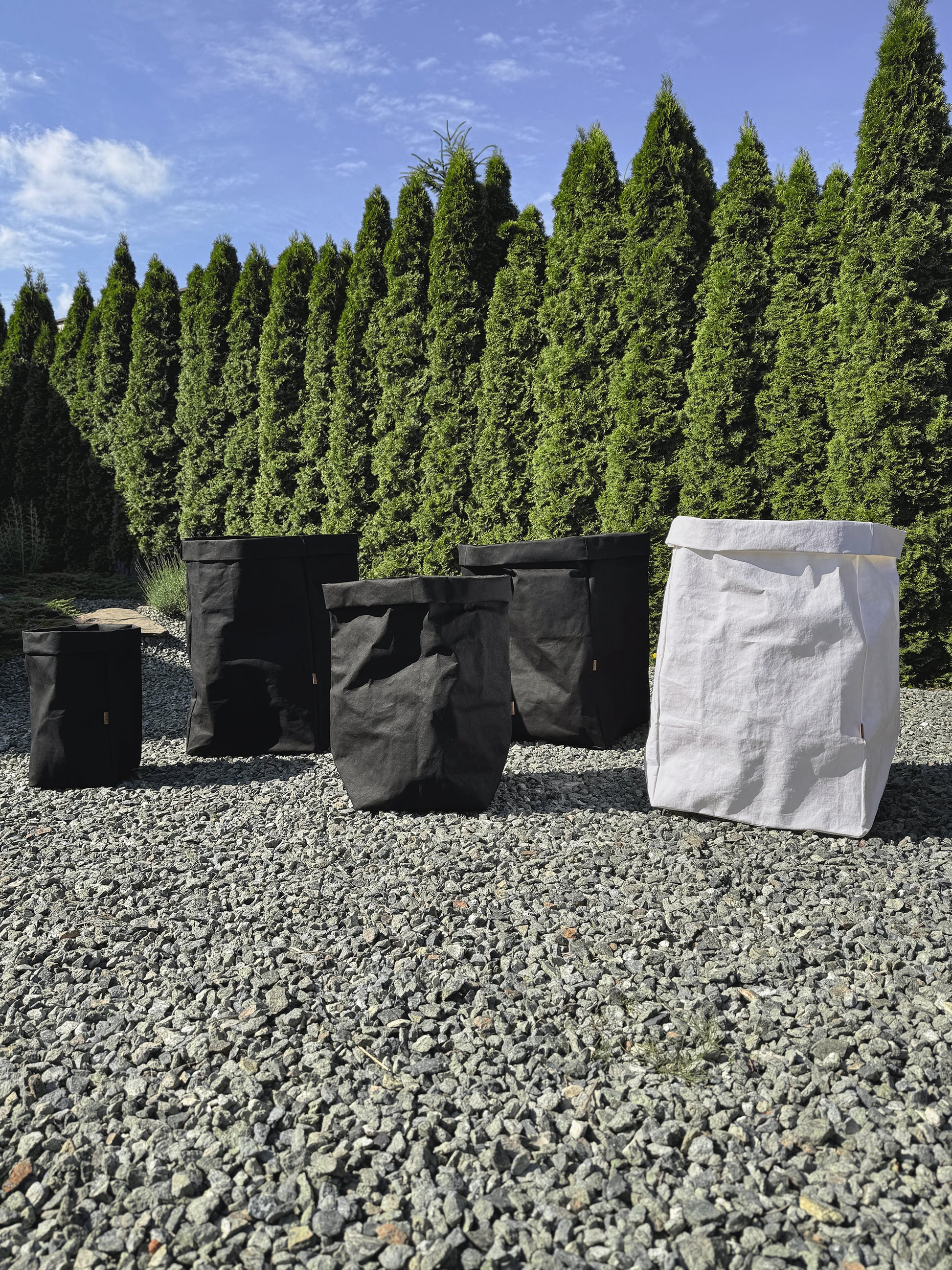 Five washable paper bags in different sizes, including four black bags and one white bag, arranged on a gravel surface with a background of tall green conifer trees and a blue sky.