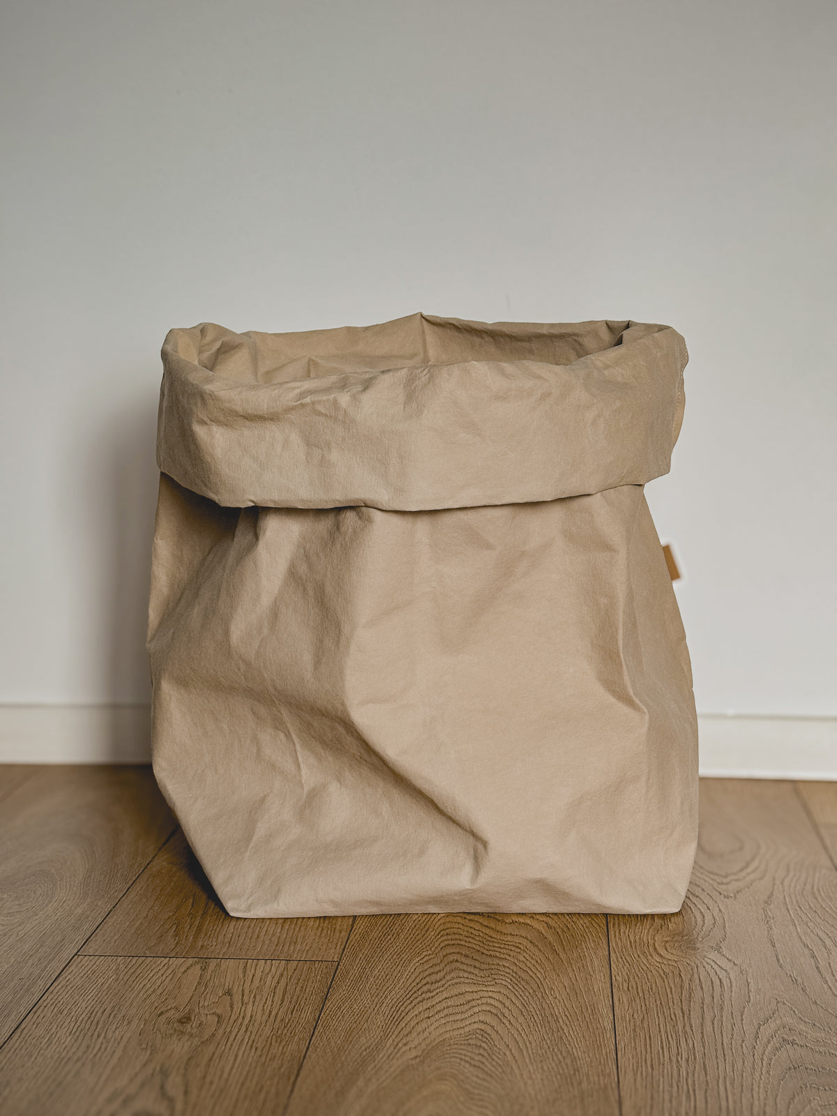 Large sand washable paper storage bag with a rolled top, wood floor against off-white wall
