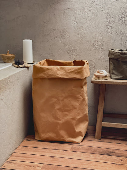 A brown washable paper laundry hamper standing on a wooden floor against a textured concrete wall, styled in a minimalist bathroom or utility room setting with a small wooden bench and soap dish nearby.