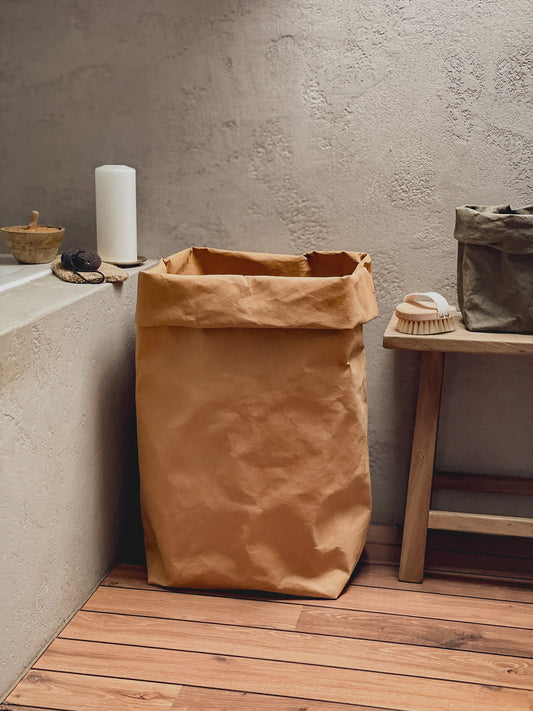 A brown washable paper laundry hamper standing on a wooden floor against a textured concrete wall, styled in a minimalist bathroom or utility room setting with a small wooden bench and soap dish nearby.